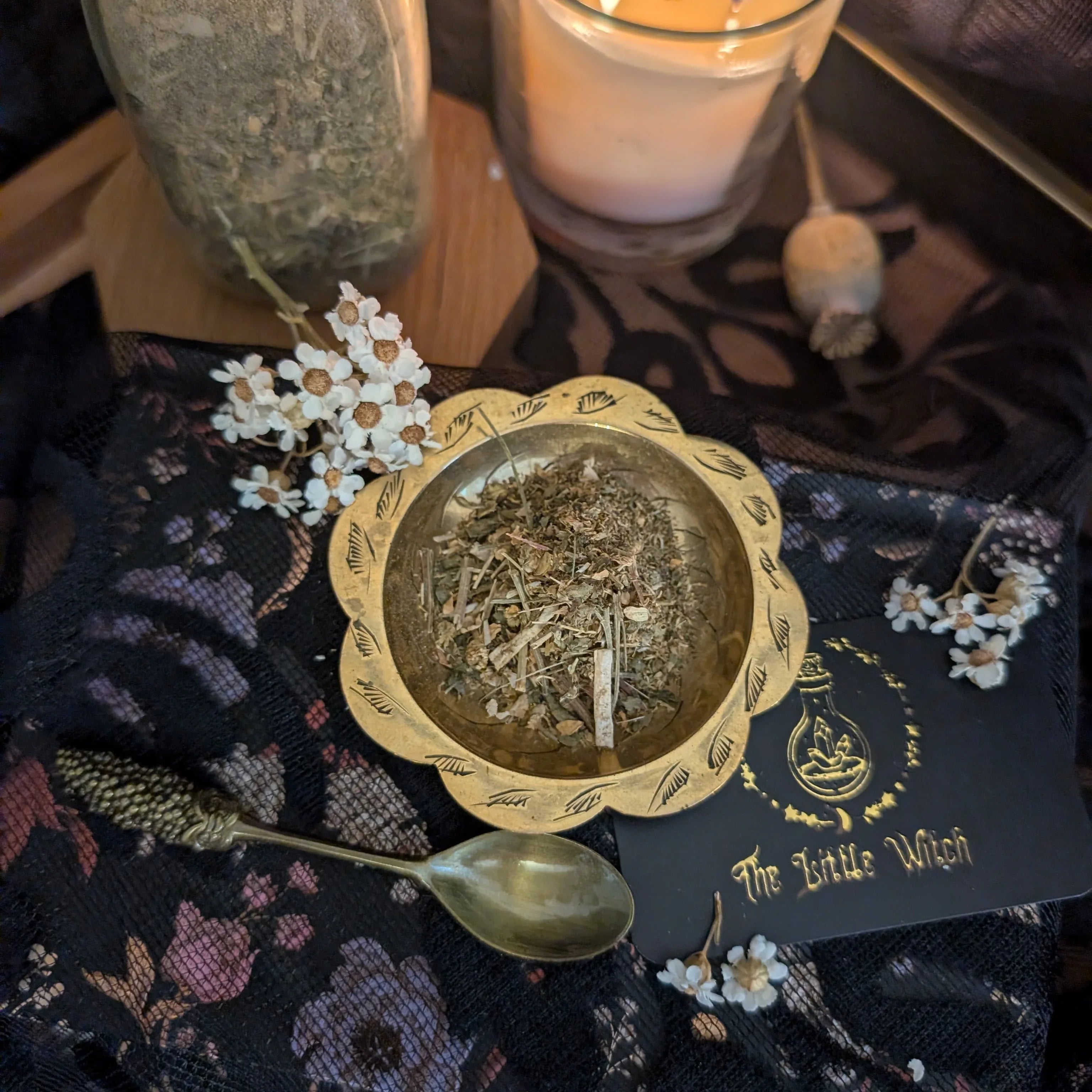 Small brass bowl with herbs on a dark fabric surface with 'The Little Witch' card.