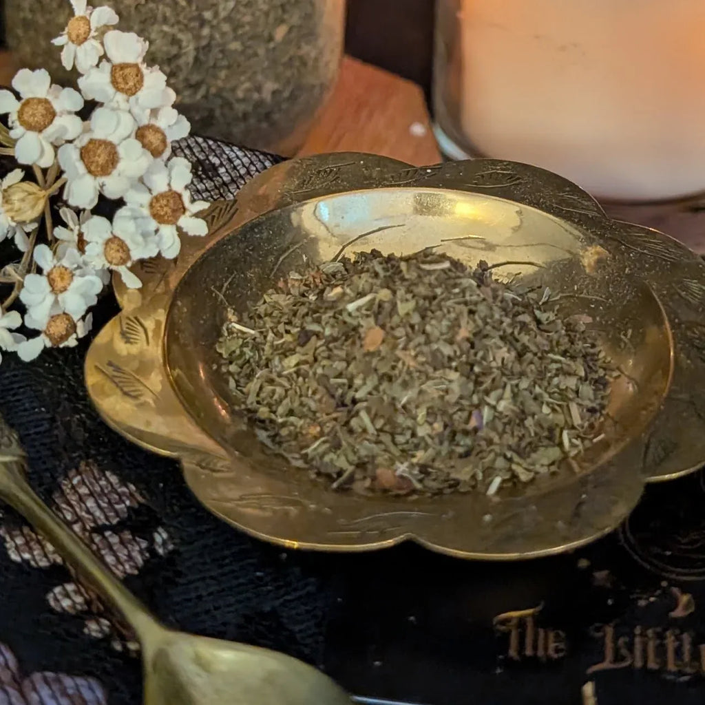 Gold bowl filled with dried  mint herbs on a textured surface with flowers and a candle in the background.