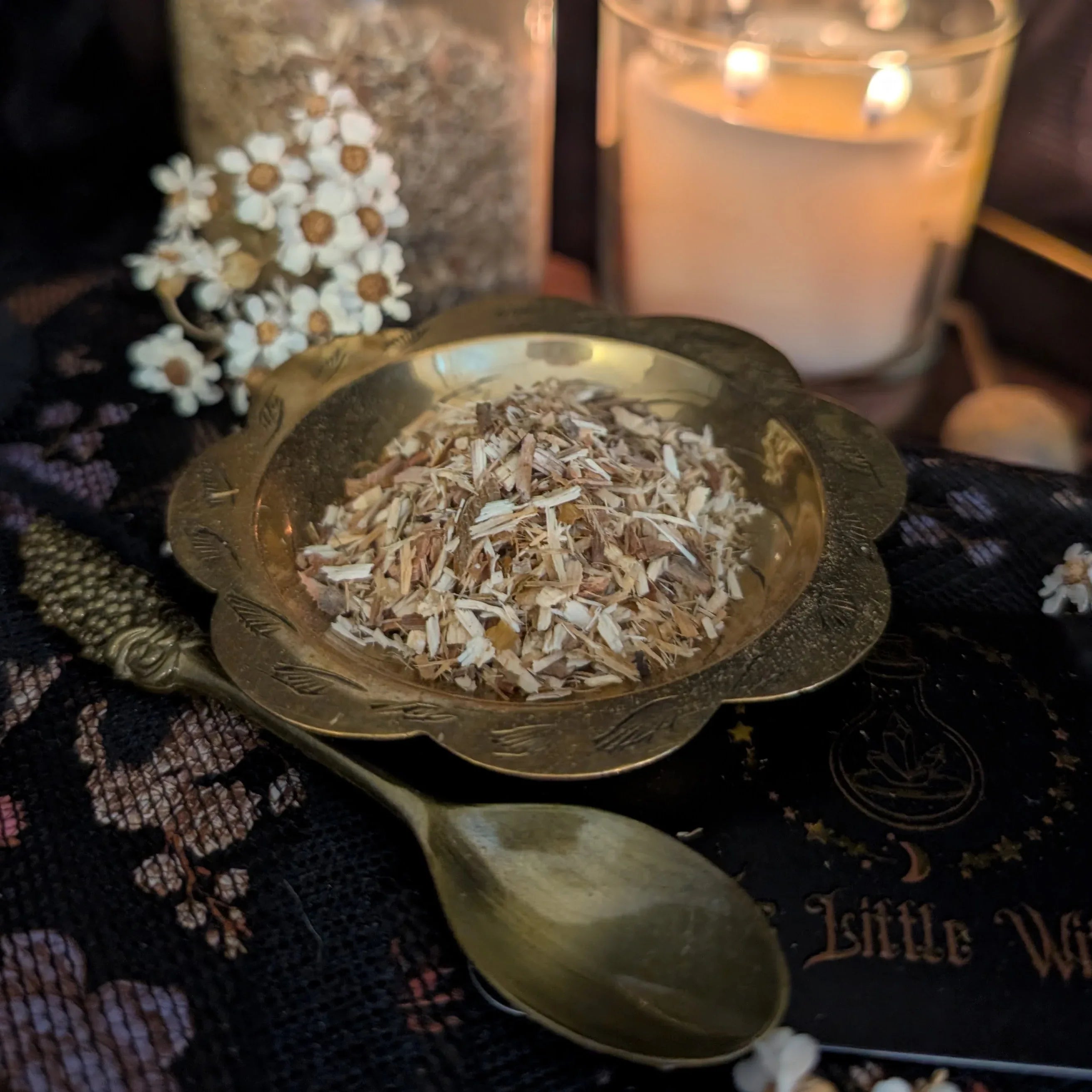 White Willow Bark herb in a brass bowl with a spoon on a dark surface, surrounded by candles and flowers.