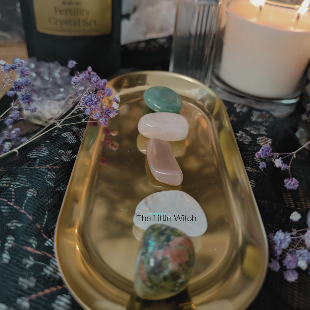 A selection of smooth crystals displayed on a gold tray with a candle and decorative flowers in the background, accompanied by a QR code and the brand name 'The Little Witch'.
