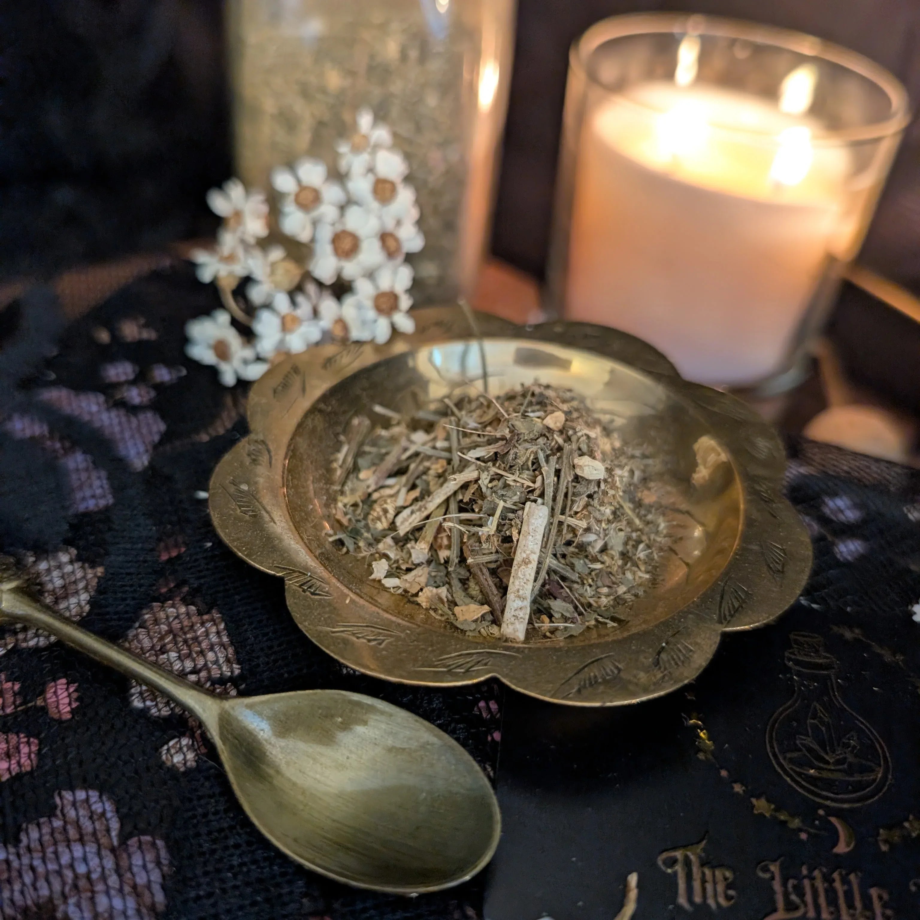 Feverfew in a brass dish with a candle and spoon on a dark fabric background