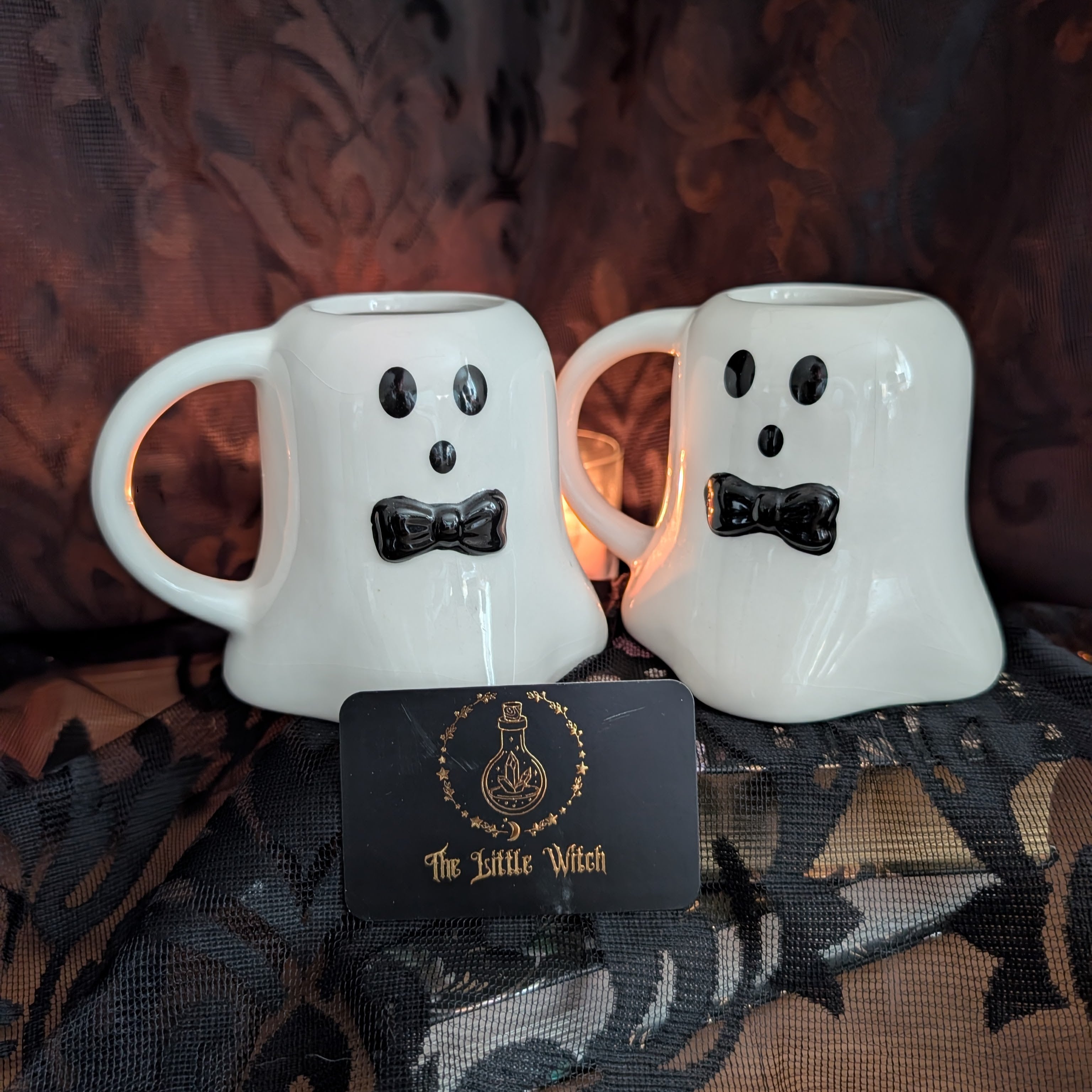 Two white ceramic mugs shaped like ghost faces with black eyes, mouths, and bow ties, displayed on a dark lace fabric backdrop. A black card with gold lettering reading 'The Little Witch' is placed in front.
