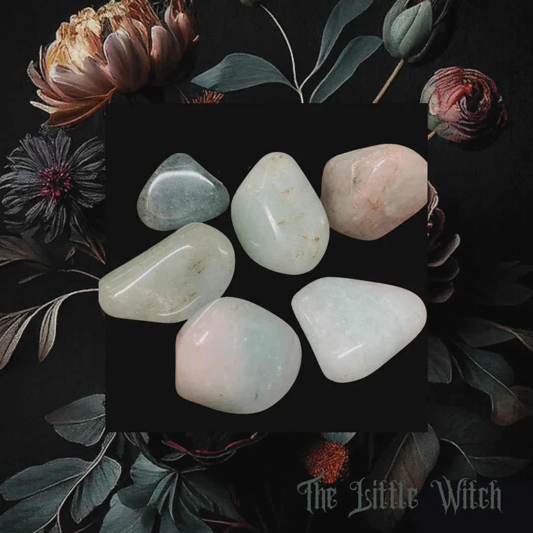 Set of tumbled stones on a dark surface with flowers and leaves, branded 'The Little Witch'.