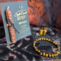 Brown spell candle with packaging and a bracelet on a textured surface