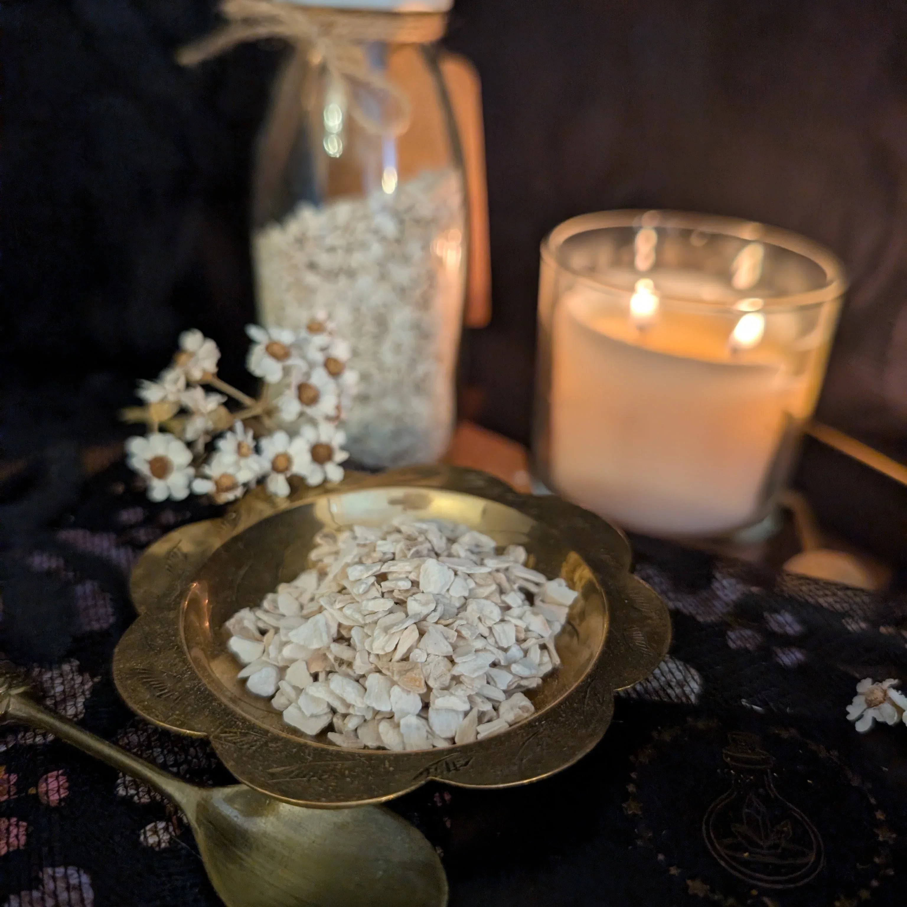 Candle, jar, and bowl of dried horseradish on a dark surface with flowers.