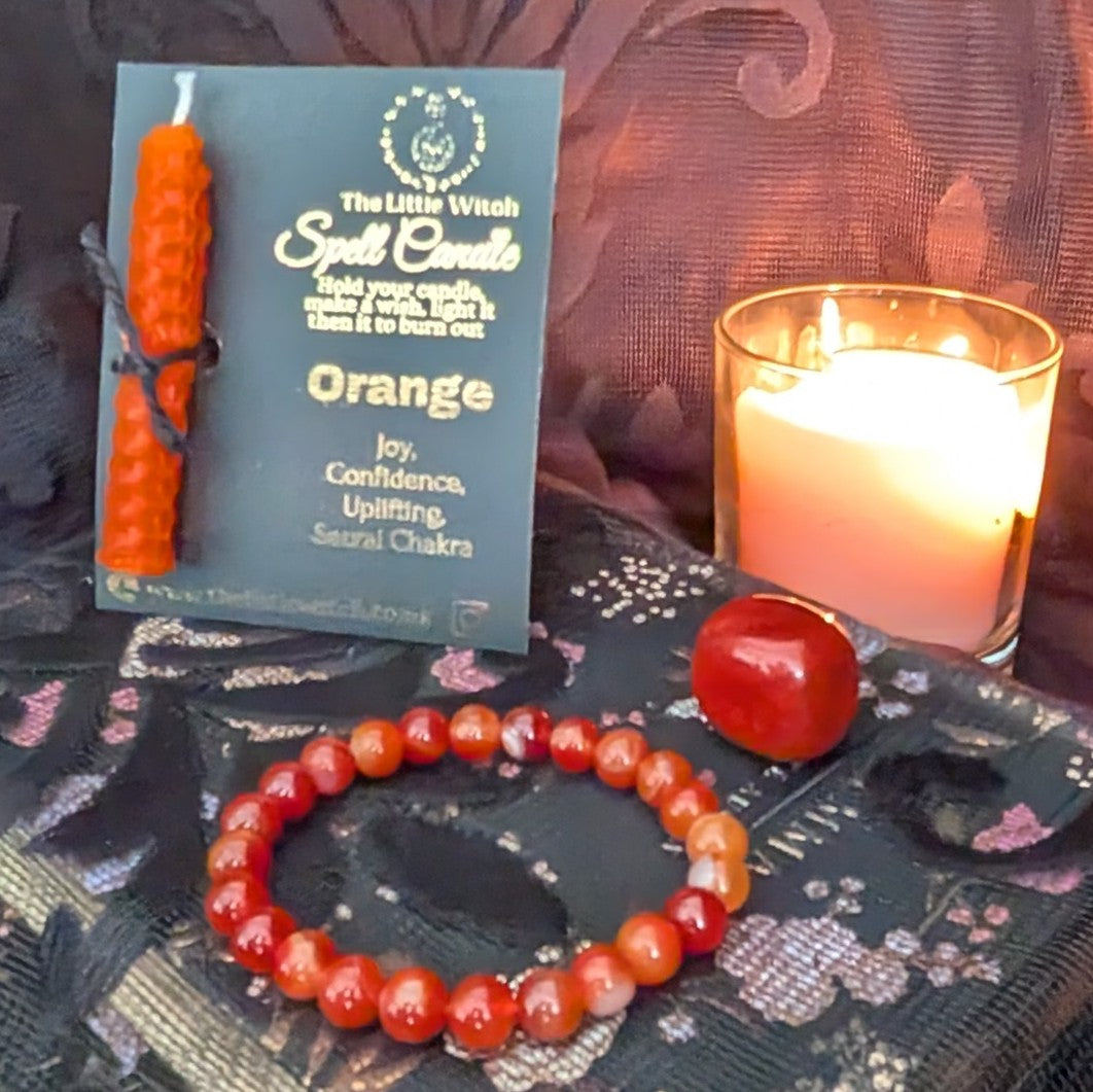 Orange spell candle with packaging, bracelet, and lit candle on a dark fabric background