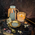 A dark moody scene with a gently flickering candlelight, a brass bowl and glass jar containing dried poppy seeds