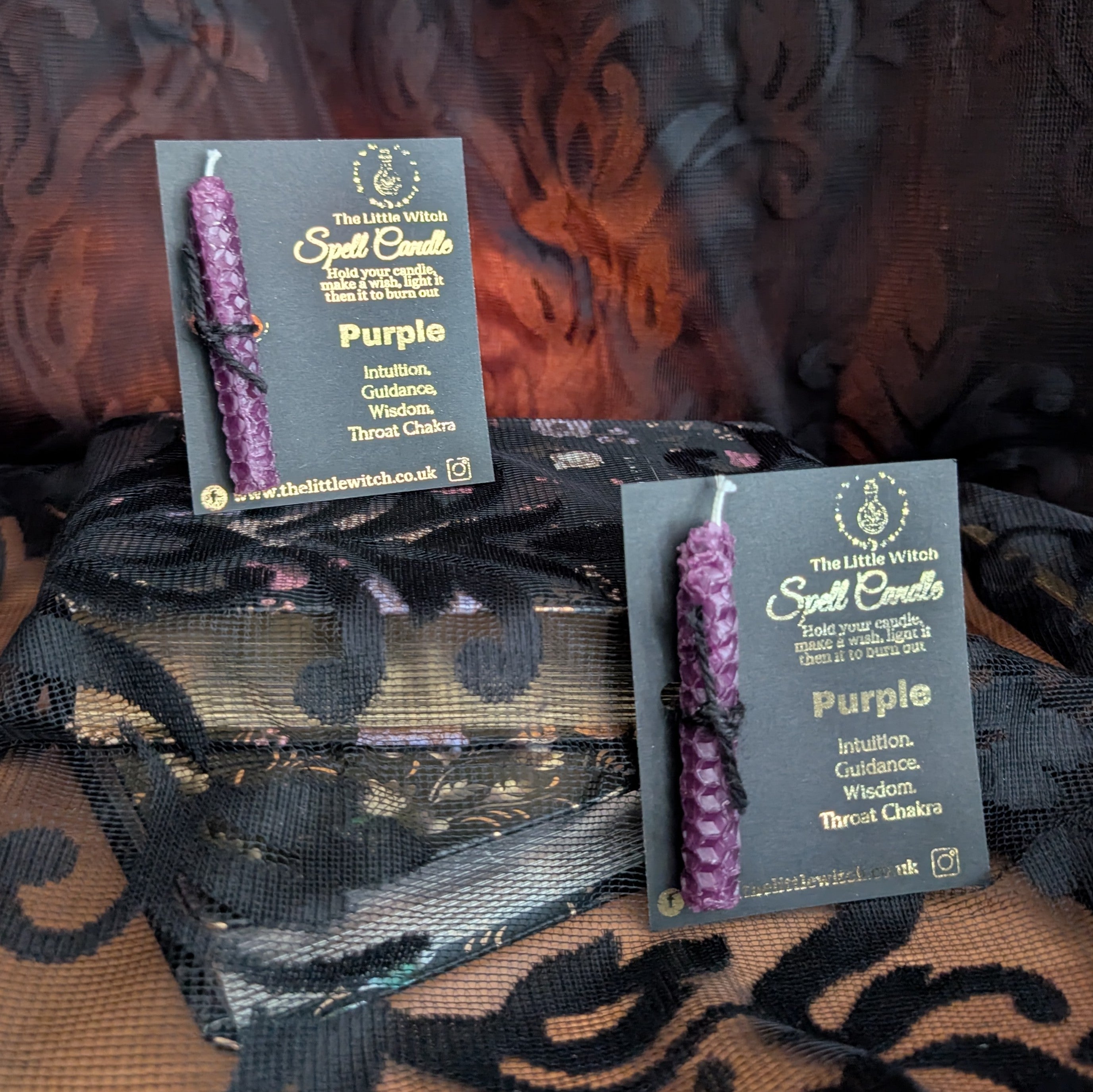 Two purple candles with The Little Witch branding on a patterned fabric background