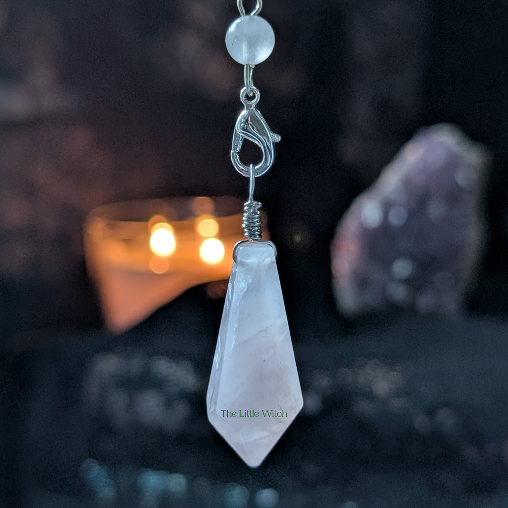 Crystal pendulum with 'The Little Witch' branding against a dark background with blurred candlelight and stones.
