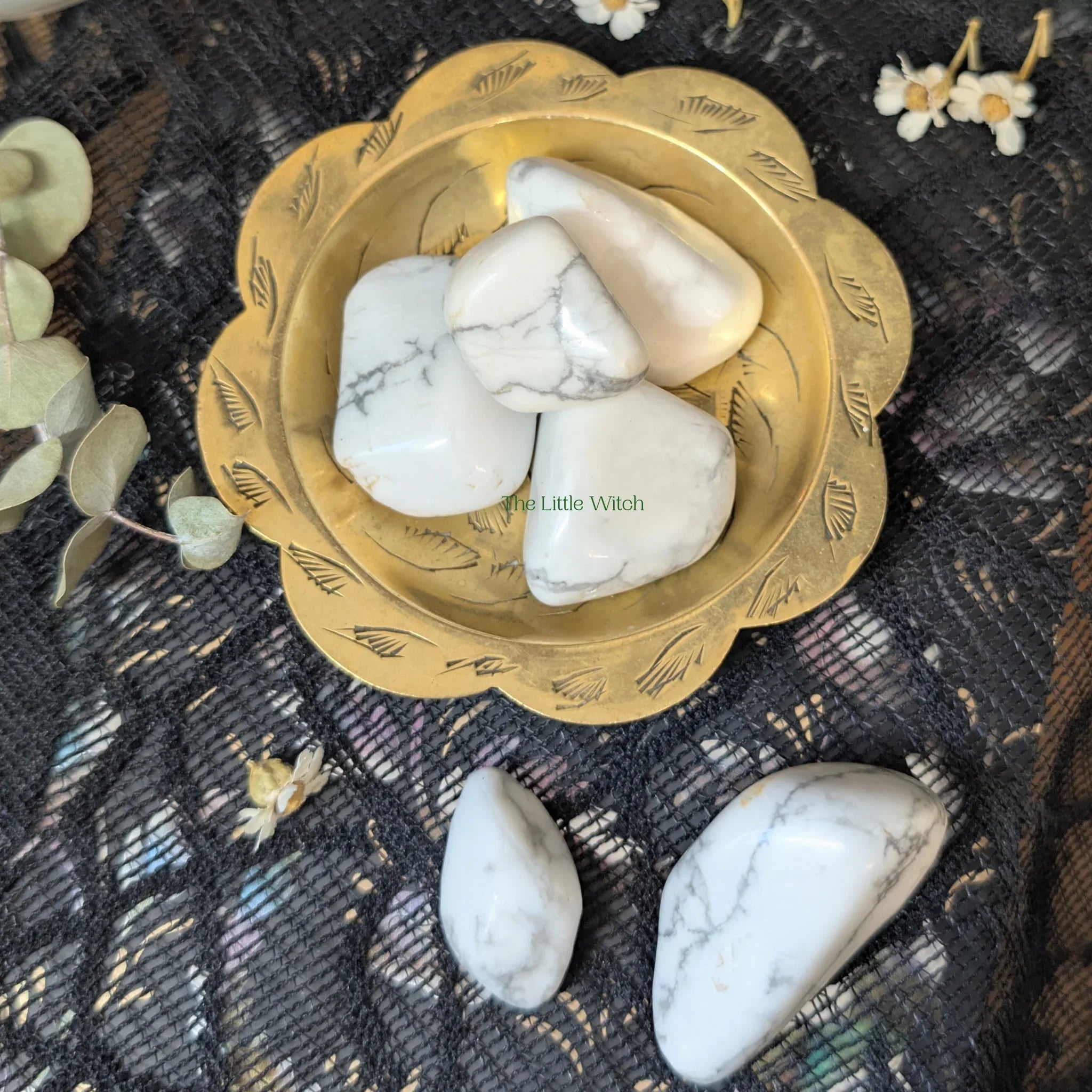 White Howlite stones in a gold bowl on a textured black surface with floral elements.