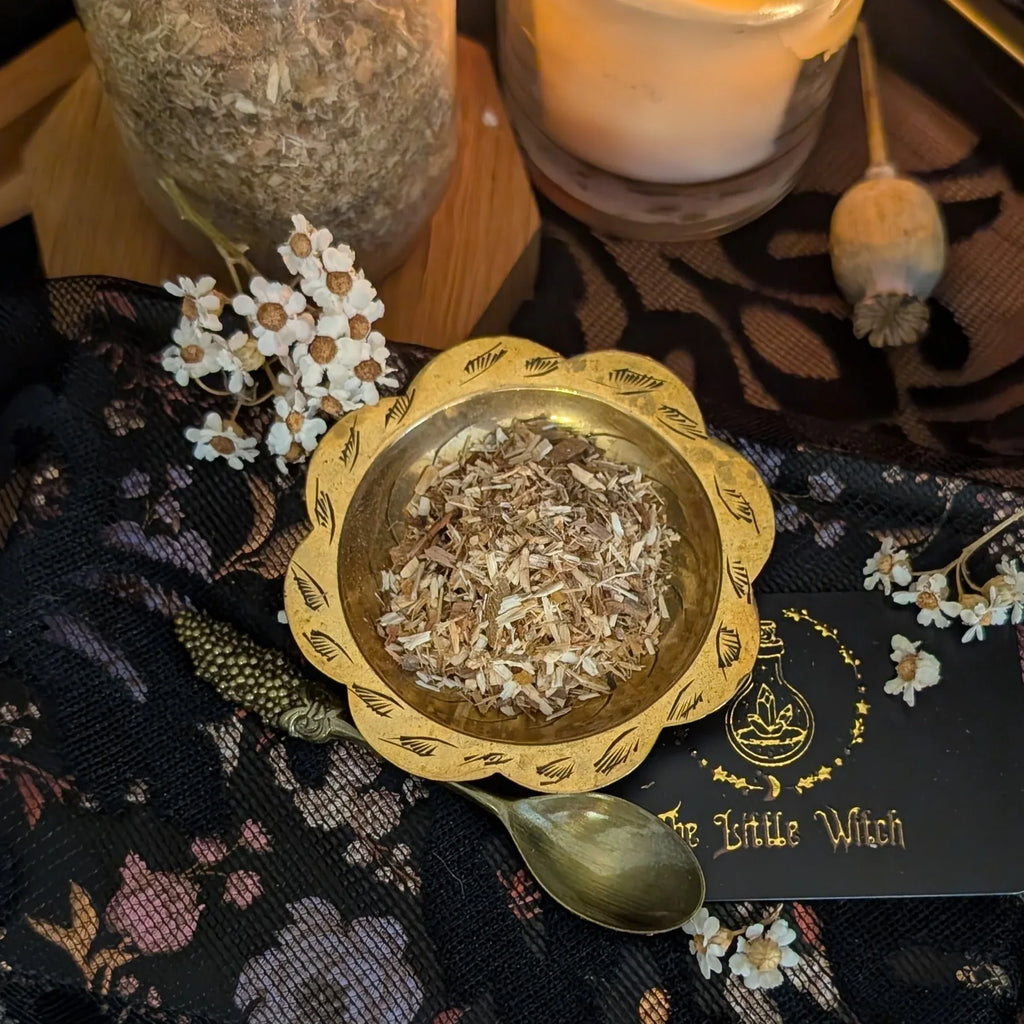 Gold bowl with dried herbs on a decorative cloth with 'The Little Witch' branding.