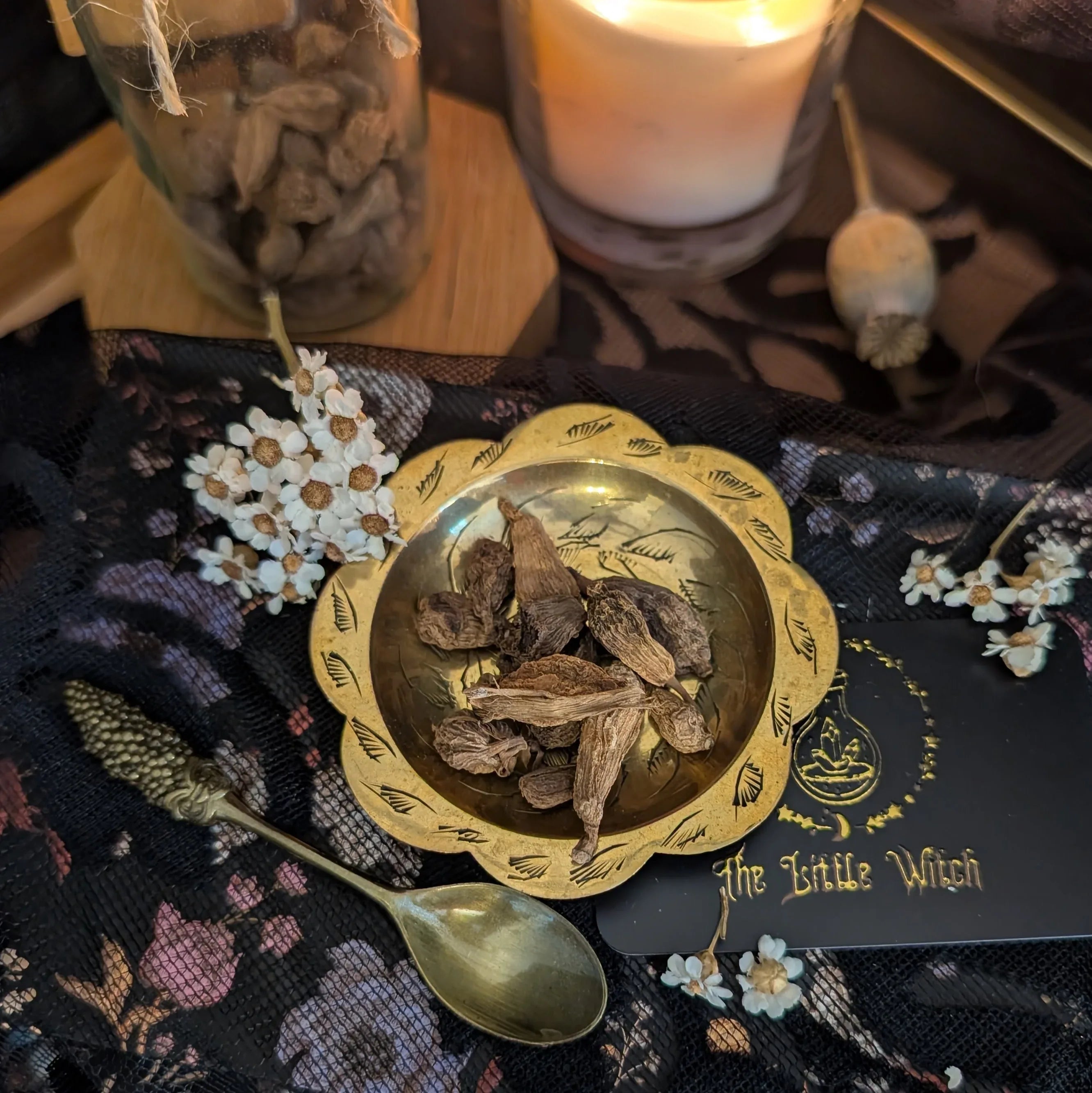 Gold dish with dried roots on a dark fabric background with flowers and a candle.