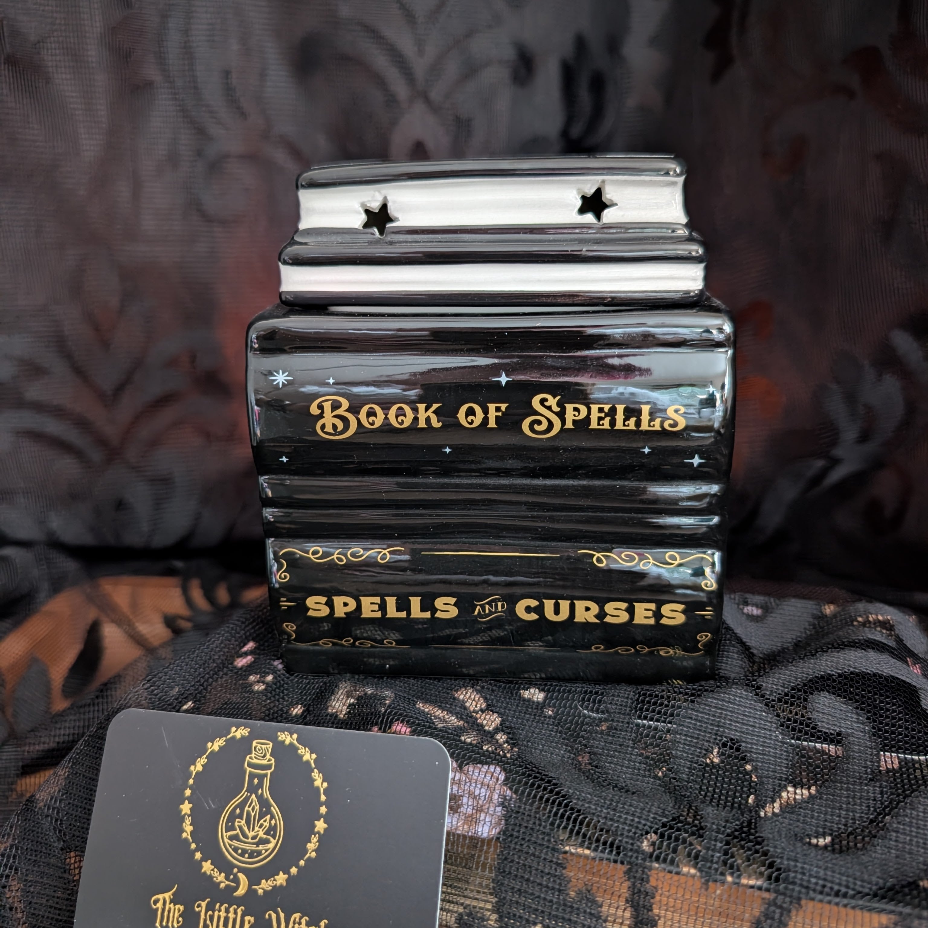 A black decorative book-shaped object labeled 'Book of Spells' and 'Spells and Curses' with gold lettering, shown on a dark patterned fabric background. A small card is partially visible in the lower left corner.